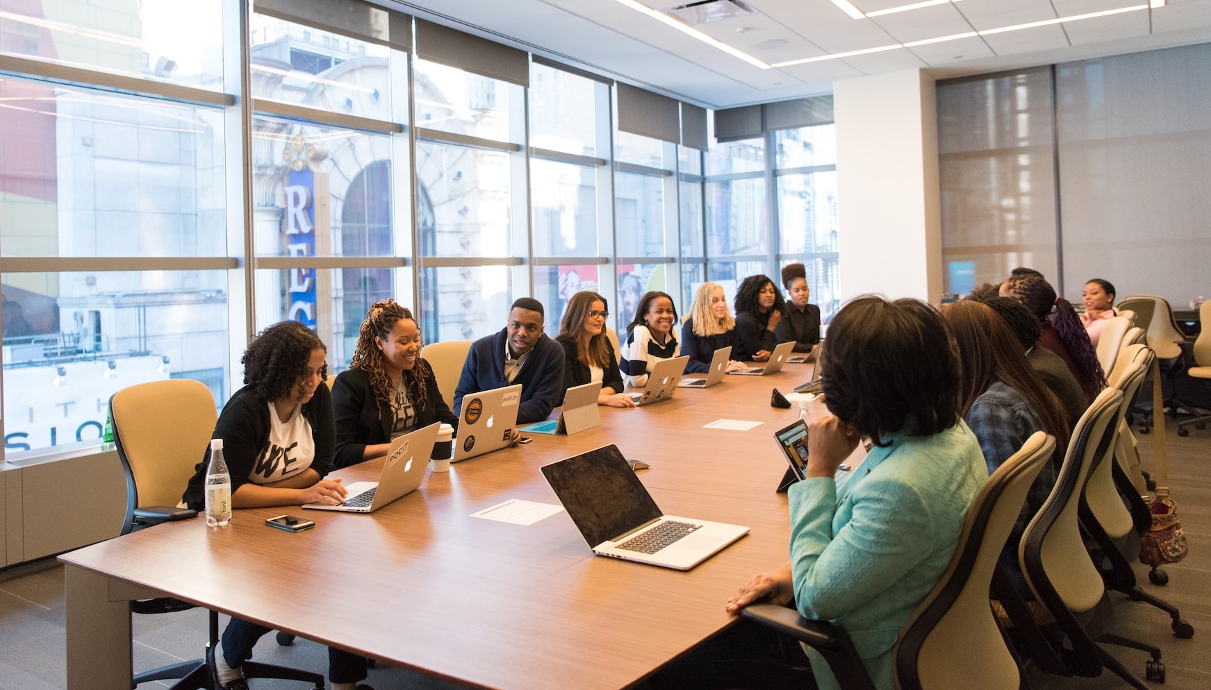 A diverse group of people sits around a large table, engaged in a collaborative meeting. They are actively discussing ideas, with notebooks and laptops open in front of them. Some participants are pointing at charts and documents, while others are listening attentively and taking notes.