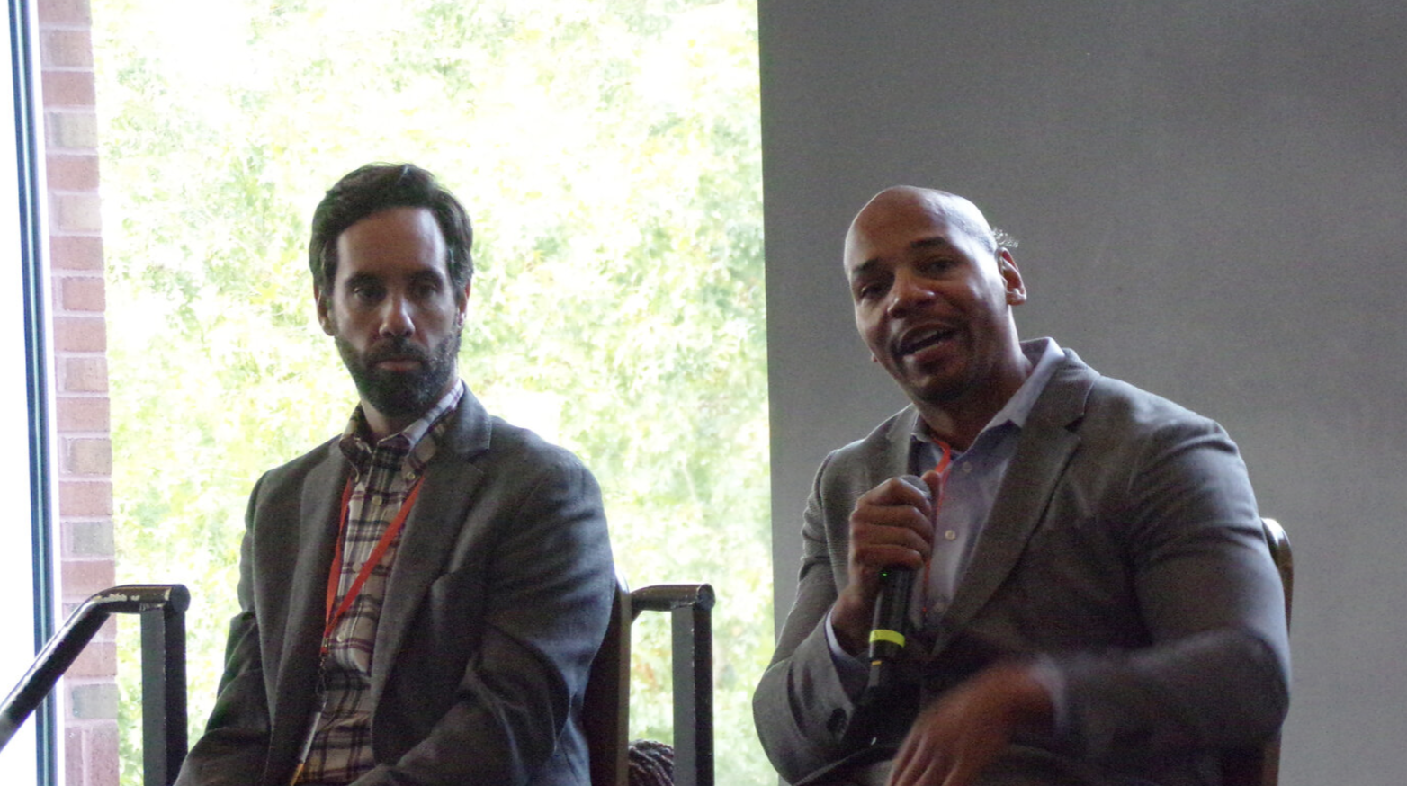 Two men sitting in chairs. One man is holding a microphone and speaking.