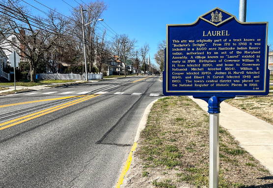 A blue sign on the side of a highway. The sign lists the history of Laurel, Delaware.