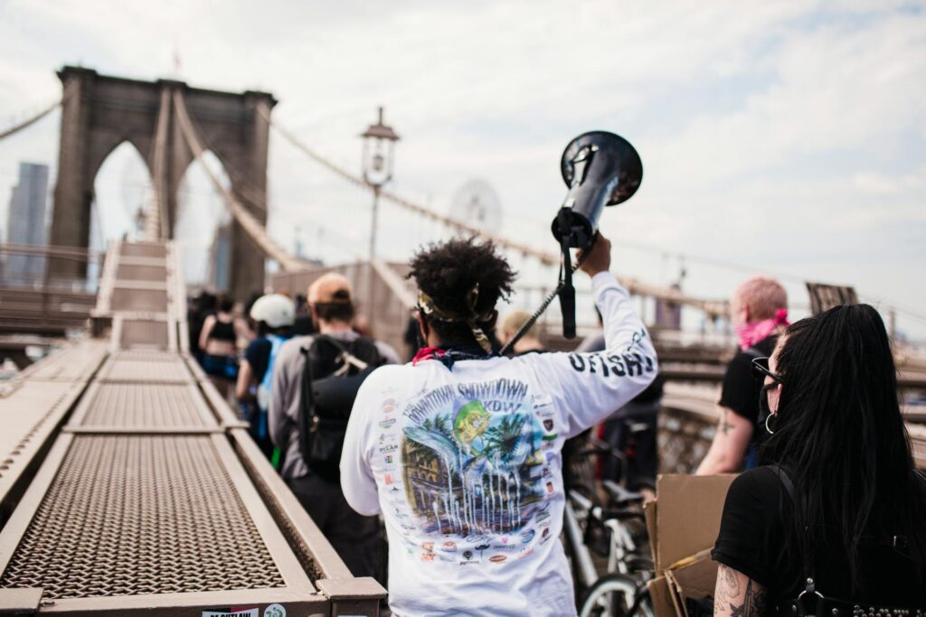 A Black person holding a megaphone aloft in their right hand and speaking into.a microphone with their left hand. Their back is to the camera, and they are on a bridge with a large number of people.